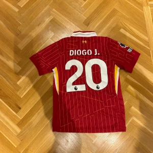 Liverpool Diogo Jota matchtröja Nike S - Liverpool FC matchtröja med Diogo Jota och nummer 20 på ryggen. Röd med gula och vita detaljer, tunna gula linjer i mönstret, klassisk krage och korta ärmar. Broderad Nike-logga och LFC-märke, tillverkad i Dri-FIT-material för bästa komfort.