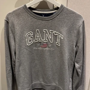 Gant Sweatshirt - Snygg grå sweatshirt från GANT med broderad logga och amerikansk flagga framtill. Klassisk rund halsringning och långa ärmar. Mjuk bomullsmix som är skön att ha på sig. Lite slitage men inga håll längst ner på ärmarna. Bra skick!
