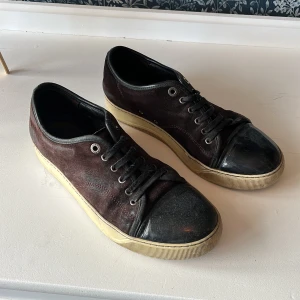 Bruna mockasneakers med svarta detaljer - Snygga sneakers i mörkbrun mocka med svarta skinnkanter och tåparti i svart blankt material. Skorna har snörning och en beige platt sula som ger en cool kontrast. Perfekta för dig som gillar en stilren men ändå unik look.