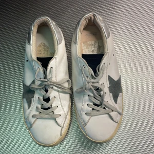 Golden Goose strl 39/40 - Snygga vita Golden Goose med grå mockastjärna på sidan och silverfärgade detaljer vid hälen. Skorna har grå snörning, platt sula och rund tå. Perfekta för dig som gillar en clean men ändå unik look. Köpta här på Plick men var tyvärr för små för mig, därav säljs dem vidare.