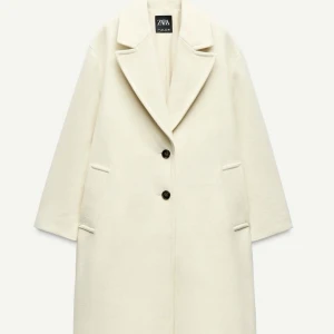 Zara crèmefärgad kappa - Säljer en stilren vit kappa från Zara i storlek XS. Kappan har klassisk krage, två knappar framtill och raka fickor. Perfekt för dig som vill ha en clean och tidlös look. Helt ny med prislappar på.