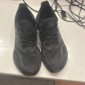 Svarta Adidas sneakers i mesh - Svarta Adidas sneakers med klassiska tre ränder på sidan. Ovandelen är i ventilerande mesh och skorna har snörning samt platt sula. Perfekta för dig som gillar en sportig och clean look. Storlek 40 2/3.