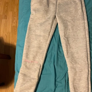 Syna Word tracksuit i färgen grå och råsa  - Snygga grå mjukisbyxor i bomull med rosa broderi 'Forgive Me' på benet och stort rosa graffiti-tryck på baksidan. Perfekta för chill dagar eller när du vill ha en avslappnad streetstyle. Mjukt material och skön passform. Det går alltid att komma överens om priset.