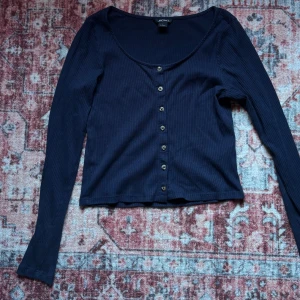 Mörkblå ribbad topp från Monki - Mörkblå ribbad topp från Monki med långa ärmar och rundad urringning. Toppen har knappar framtill och är tillverkad i mjuk bomull. Perfekt att styla med jeans eller kjol för en chill look.