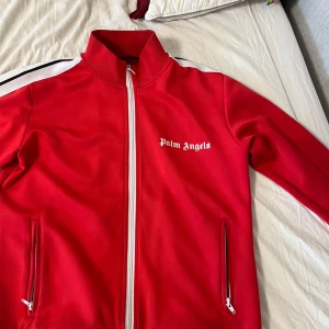 Röd Palm Angels track jacket - Röd track jacket från Palm Angels med vita och svarta ränder längs ärmarna. Jackan har hög krage, hel dragkedja framtill och två fickor med dragkedja. Materialet är mjukt och sportigt, perfekt för en streetwear-look.