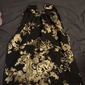 Svart och guld blommig festtopp - Snygg svart kortklänning med guldiga blommor och hög hals. Toppen  är ärmlös och har ett elegant blommigt mönster som verkligen sticker ut. Perfekt för dig som vill ha en unik och glamorös look. Materialet känns mjukt och har en lätt glans.