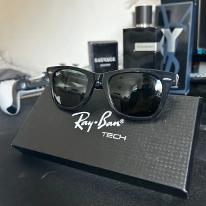 Svarta Ray-Ban Wayfarer Tech solglasögon - Säljer ett par stilrena svarta Ray-Ban Wayfarer Tech solglasögon med klassisk fyrkantig form. Bågarna är i blankt svart syntetmaterial och har Ray-Ban-logga på sidan. Perfekta för dig som vill ha en ikonisk look och skydda ögonen mot solen.