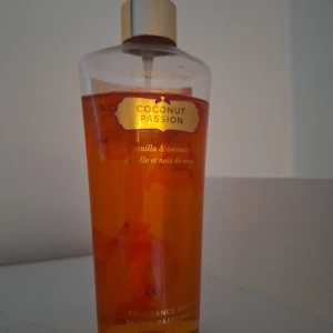 Victoria's Secret Coconut Passion mist - Fräsch och tropisk body mist från Victoria's Secret med doft av vanilj och kokos. Flaskan är genomskinlig med gyllene lock och etikett, och innehållet har en varm orange ton. Perfekt för dig som älskar söta och exotiska dofter.