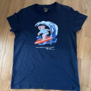 Mörkblå Polo Bear t-shirt från Ralph Lauren -  Mörkblå t-shirt från Polo Ralph Lauren med ikoniskt Polo Bear-tryck där björnen surfar på en våg. Klassisk rund hals och korta ärmar. Tillverkad i mjuk bomull, perfekt för en chill och stilren look. M men passar S