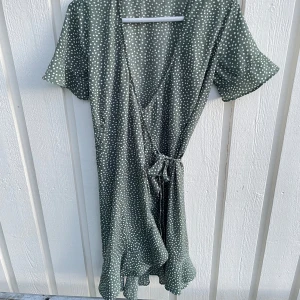 Grön prickig omlottklänning med knyt - Snygg grön kort omlottklänning med vita prickar och knyt i midjan. Klänningen har v-ringad hals, korta ärmar och volangkant nedtill. Perfekt för dig som gillar en chill och trendig stil. Materialet känns lätt och skönt mot huden.