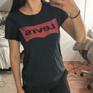 Svart Levi's t-shirt med röd logga - Svart t-shirt från Levi's i mjuk bomull med klassisk röd logga framtill. Modellen är kortärmad och har rund hals. Perfekt för dig som gillar streetstyle och vill ha en ikonisk look med tydlig branding.
