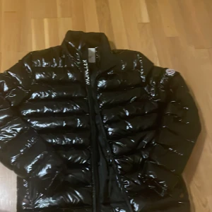 Svart pufferjacka från Moncler L - Säljer en svart, glansig pufferjacka från Moncler i storlek L. Jackan har klassisk quiltad design, dragkedja framtill och Moncler-logga på ärmen. Insidan har en röd detalj och ett coolt serietidningsmärke. Perfekt för kalla dagar och riktigt snygg streetstil.Kan också diskutera om pris det är bara att skriva.
