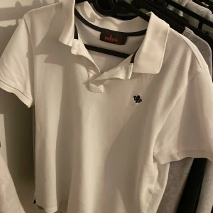 Vit pikétröja från Morris - Fräsch vit pikétröja från Morris med klassisk krage och korta ärmar. Tröjan har en liten broderad logga på bröstet och är gjord i mjuk bomull som andas. Perfekt för en clean och stilren look.