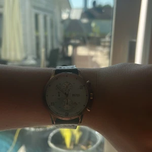 Silverfärgad BOSS-klocka med brun läderrem - Stilren herrklocka från BOSS med rund silverfärgad urtavla och tre mindre visartavlor. Klockan har en klassisk brun läderrem med snygga sömmar och metallspänne. Kronograf-funktion och tydliga siffror på urtavlan ger en modern och tidlös look. Lite sliten ovanför klockan som man ser på bilderna. Ny pris: 1500kr-2000kr. Nummer har lossnat 