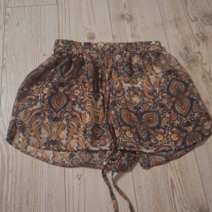 Strand shorts - Säljer ett par superlätta och luftiga strand shorts i beige, orange och blått. De har elastisk midja och snörning framtill. Perfekta för varma sommardagar och riktigt sköna att ha på sig över bikinin. Materialet känns tunt och svalt.