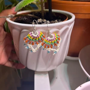 Färgglada handgjorda makraméörhängen - Unika örhängen i makramé med regnbågsfärgade trådar i lila, gul, grön, orange och vitt. Dekorerade med små blå pärlor längst ner för extra detalj. Perfekta för dig som vill sticka ut med boho-vibe och färg. Säljs som par.