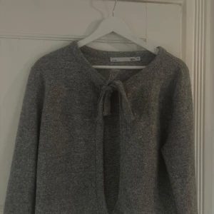 Grå kofta med knyt från 157 -  grå kofta från 157 som man kan knyta till en rosett framtill. Koftan har lång ärm och är tillverkad i ett mjukt, stickat material som passar perfekt för kyligare dagar. Enkel och stilren design som är lätt att matcha med olika outfits🩶