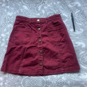 Vinröd kortkjol från Monki - Vinröd kortkjol från Monki i 100% bomull. Kjolen har två stora fickor framtill, knappar hela vägen ner och bälteshällor i midjan. Snygg A-linjeformad passform som ger en chill vibe.