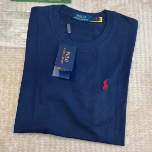 Mörkblå t-shirt från Polo Ralph Lauren - Snygg mörkblå t-shirt från Polo Ralph Lauren i mjuk bomull. Klassisk rund hals och korta ärmar. På bröstet finns det ikoniska röda Polo-logot broderat. Perfekt basic med stilren vibe och skön passform. Mått: 63cm lång x 46cm bred.