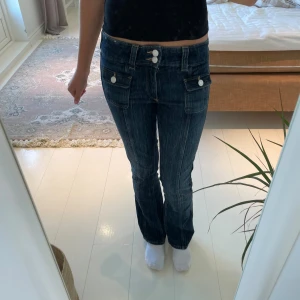 Mörkblå bootcut jeans med fickdetaljer - Snygga mörkblå jeans med bootcut passform och vita knappar på fickorna framtill. Jeansen har låg midja och markerade sömmar som ger en cool look. Perfekta för dig som gillar klassiska jeans med en twist.