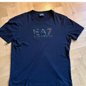 Svart EA7 Emporio Armani t-shirt - Snygg svart t-shirt från EA7 Emporio Armani med stort glansigt logotryck på bröstet. Klassisk rund halsringning och korta ärmar. Tillverkad i mjuk bomull som känns skön mot huden. Perfekt för dig som gillar stilrena och exklusiva plagg.