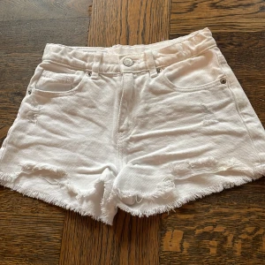 Vita jeansshorts med fransar från Zara - Snygga vita jeansshorts från Zara med fransiga kanter och slitna detaljer. Modellen är högmidjad med klassiska fem fickor, knapp och dragkedja framtill. Perfekt för dig som gillar en cool och avslappnad stil på sommaren.