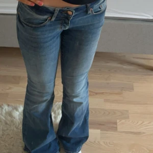Blå bootcut jeans med låg midja - Snygga blå jeans med bootcut och låg midja. Jeansen har klassisk femficksdesign, dragkedja och knapp framtill. Materialet är stretchigt denim som sitter skönt och formar sig efter kroppen. Perfekta för dig som gillar en retroinspirerad look.