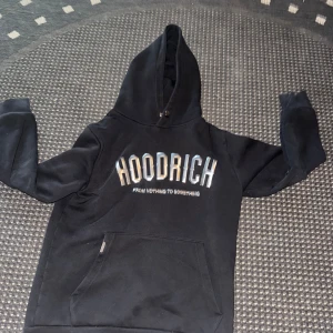 Svart HOODRICH hoodie med tryck - Svart hoodie från HOODRICH med stor logga och texten 'FROM NOTHING TO SOMETHING' på bröstet. Klassisk känguruficka framtill och huva med snörning. Perfekt för en chill och street-inspirerad stil.