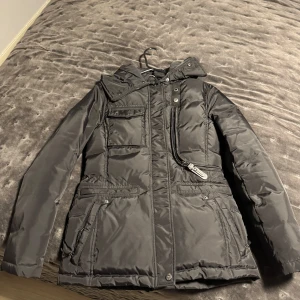 Svart pufferjacka från Hollies - Snygg svart pufferjacka från Hollies med huva och flera praktiska fickor. Jackan har dragkedja och knappar framtill samt coola detaljer som snedställda fickor och markerade sömmar. Perfekt för kalla dagar när du vill hålla stilen.