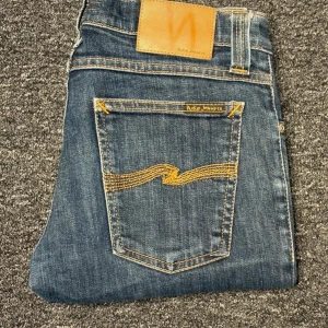 Nudie Jeans blå jeans W28 L34 - Snygga blå jeans från Nudie Jeans med klassisk gul kontrastsöm och ikonisk broderad bakficka. Modellen har normal passform och raka ben. Tillverkade i slitstarkt jeanstyg med detaljerade sömmar och läderpatch bak i midjan.
