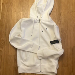 Vit hoodie från Polo Ralph Lauren - Vit hoodie från Polo Ralph Lauren med dragkedja framtill, huva med snörning och klassisk logga broderad på bröstet. Tillverkad i mjuk bomullsblandning, perfekt för en clean och stilren look. Långa ärmar och fickor framtill.