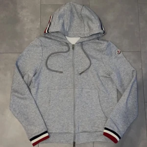 Grå Moncler hoodie med dragkedja - Snygg grå hoodie från Moncler med dragkedja, huva och snörning. Ärmsluten har röd, vit och marinblå rand. Moncler-logga på ena ärmen. Mjuk bomullskänsla, perfekt för chill dagar eller när du vill ha en clean look.