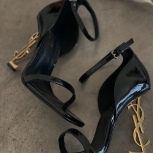 Svarta Saint Laurent klackar med YSL-häl - Säljer ett par svarta Saint Laurent klackar i lackat skinn med ikonisk guldig YSL-logga som klack. Skorna har smal rem över tårna, justerbar ankelrem och fyrkantig tå. Riktigt statement piece för dig som vill sticka ut.