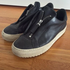 Arigato med dragkedja - Stilrena svarta sneakers i skinn från Axel arigato med chunky vit sula och cool dragkedjedetalj framtill istället för klassisk snörning. Låg modell med rund tå och räfflad undersida för extra grepp. Perfekta för dig som gillar minimalistisk och modern stil.