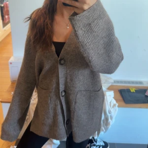 Brun stickad kofta med knappar - Mysig brun stickad kofta med stora knappar framtill och två rejäla fickor. Koftan har en loose passform och långa ärmar, perfekt att slänga på sig över en t-shirt eller linne. Enkel och stilren design som funkar till det mesta.