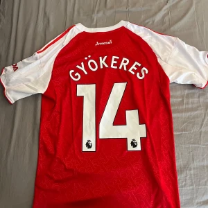 Arsenal Gyökeres 25/26 matchtröja - Säljer en  Arsenal fotbollströja från Adidas med Gyökeres och nummer 14 på ryggen. Tröjan är röd med vita korta ärmar, vita detaljer och klubbmärke på bröstet. Materialet är lätt och andas, perfekt för match eller träning. Snygga mönster i tyget och klassisk rund hals .