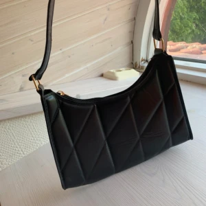 Svart quiltad axelväska - Snygg svart axelväska med quiltad design och guldfärgade detaljer. Väskan har en stilren form och dragkedja upptill. Perfekt för att lyfta din outfit med en trendig touch.