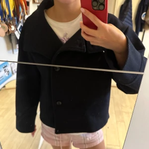 Svart skepparkavaj i ull - Säljer en svart skepparkavaj med klassisk krage och stora knappar framtill. Jackan är i ullmaterial och har en boxig, lite oversized passform med långa ärmar. Perfekt för dig som vill ha en stilren och tidlös look.