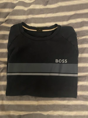 BOSS Sweatshirt - Snygg svart sweatshirt från BOSS i storlek M. Tröjan har en blå rand över bröstet med BOSS-loggan tryckt på vänster sida. Rund hals och långa ärmar, perfekt för chill dagar. Materialet är mjuk bomull som känns skönt mot huden.
