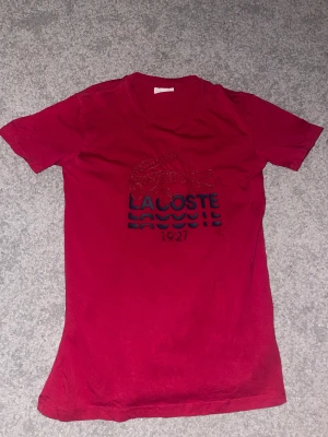 Röd Lacoste t-shirt med tryck - Snygg röd t-shirt från Lacoste med coolt krokodiltryck och texten 'Lacoste 1927' på bröstet. Klassisk rund hals och korta ärmar. Materialet är mjuk bomull som känns skönt mot huden. Perfekt för dig som gillar streetwear och sportig stil.