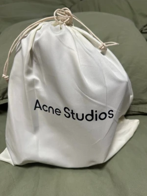 Svart handväska Acne Studios skinn - Minimalistisk svart handväska från Acne Studios i slätt skinn. Väskan har en unik form med knutdetalj på sidorna och robusta handtag. Kommer med original dustbag i vitt tyg. Perfekt för dig som gillar stilren och modern design.