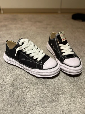 Mihara Yasuhiro Sneakers - Unika svarta sneakers från MMY med grov vit platt sula och vit tå. Skorna har kraftiga vita snören, silverfärgade öljetter och synliga vita sömmar. Ovandelen är i canvas och har en cool, avslappnad vibe. Perfekt för dig som gillar streetstyle och vill sticka ut. 