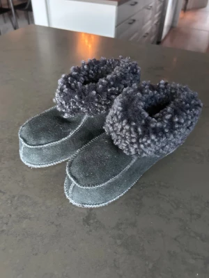 Handgjorda fårskinnstofflor  med ullkant till barn  - Mysiga grå handgjorda tofflor i fårskinn med fluffig ullkant runt ankeln. De har en mjuk, platt sula och är perfekta för att hålla fötterna varma hemma. Tofflorna passar barn med skostorlek ca 30-31 i skostorlek gissningsvis. Sulans yttermått är 22 cm. 