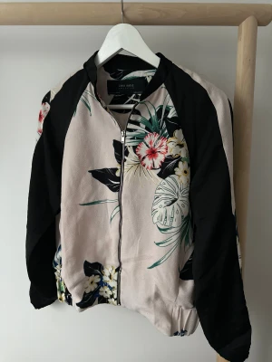 Blommig bomberjacka från Zara  - Säljer en snygg bomberjacka från Zara Basic med blommigt mönster i svart, beige, grönt och inslag av rött och blått. Jackan har svarta ärmar, dragkedja framtill och elastisk midja.
