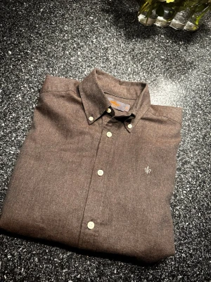 Brun skjorta från Morris - Snygg brun skjorta från Morris med klassisk krage och vita knappar. På bröstet finns ett stilrent broderat emblem i vitt. Skjortan är gjord i mjuk bomull och har en clean, normal passform som funkar till många stilar. Storlek XS men passar mer som S. Nypris 1500 kr