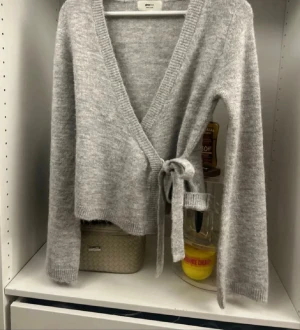 Grå omlottkofta med knyt - Mysig grå omlottkofta från Gina Tricot med långa ärmar och knyt i sidan. Koftan har ribbade kanter och en v-ringad hals, perfekt för lager på lager. Materialet är mjukt och stickat, vilket gör den extra skön att bära.