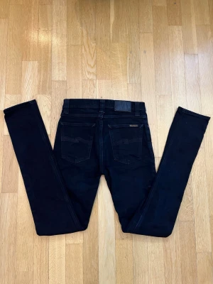 Svarta  jeans från Nudie Jeans - Säljer ett par svarta jeans från Nudie Jeans i storlek W27 L32. Jeansen har klassisk femficksdesign, svarta sömmar och Nudie Jeans-logga på bakfickan. Materialet är mjukt och stretchigt denim för en bekväm passform.