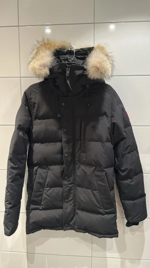Svart Canada Goose dunjacka med päls - Svart dunjacka från Canada Goose med fluffig naturfärgad päls på huvan, ikonisk röd logotyp på ärmen och flera praktiska fickor. Jackan har dragkedja och knappar framtill samt justerbar huva med snörning. Perfekt för kalla vinterdagar.