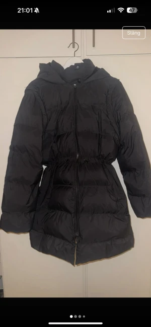 Svart pufferjacka från Primark Maternity - Svart pufferjacka från Primark Maternity i storlek M. Jackan har huva, dragkedja och snörning i midjan för en snygg siluett. Insidan är beige och materialet är syntet. Perfekt för kalla dagar med sin långa modell och quiltade design. Går även att ta av armarna och göra de till en väst.