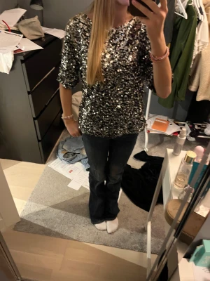 Glittrig paljettopp från H&M - Säljer en svart topp från H&M täckt med silvriga paljetter. Modellen är rak med rund halsringning och korta ärmar. Perfekt för dig som vill sticka ut med en glittrig look under kanske en fest, nyår eller jul. Materialet är polyester och toppen har en cool, festlig vibe. Köpt för 300kr. Använd 1-2 gånger